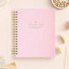 A light pink spiral-bound notebook with gold wire binding and gold foil text reading 'DAY DESIGNER.' on the cover, styled on a pale pink surface with decorative dried grass, a small dish of beads, and paper notes around it.