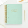 A mint green spiral-bound planner or notebook with gold wire binding and gold foil text on the cover, styled on a white surface with a gold pen nearby.