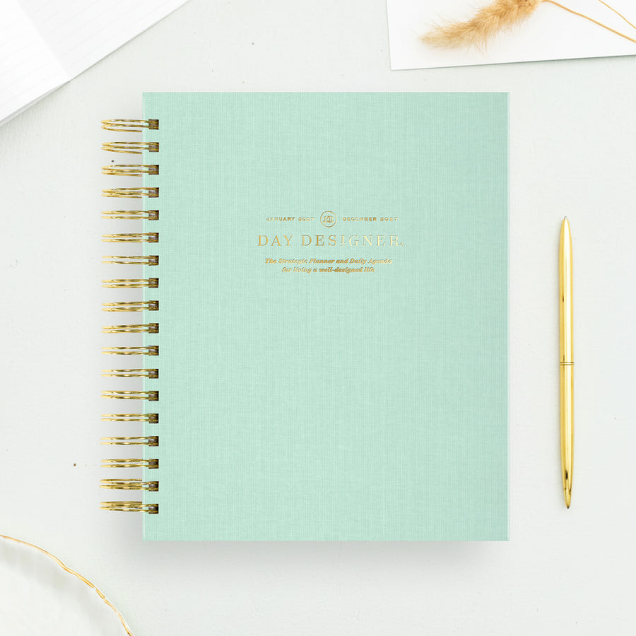A mint green spiral-bound planner or notebook with gold wire binding and gold foil text on the cover, styled on a white surface with a gold pen nearby.