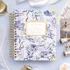 A spiral-bound planner or notebook with a white cover decorated in blue floral and swan illustrations, featuring a gold-bordered label that reads 'DAY DESIGNER.' The item is styled in a flat lay on a pale blue surface with decorative props including a feather, brass telescope, perfume bottle, and printed card.