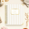 A spiral-bound planner or notebook titled 'DAY DESIGNER' lies on a light cream surface with decorative props around it. The cover has thin vertical gold stripes on a white background and a gold label in the center.