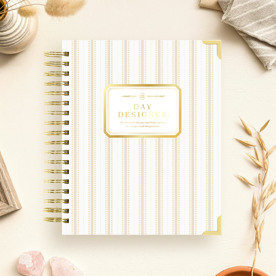 A spiral-bound planner or notebook titled 'DAY DESIGNER' lies on a light cream surface with decorative props around it. The cover has thin vertical gold stripes on a white background and a gold label in the center.