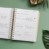mini weekly planner, open book, written on pages, gold binding and tabs, green background, palm leaves, brown dish