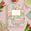 A spiral-bound planner or notebook titled 'DAY DESIGNER.' lies on a pink surface, surrounded by tulips, a green striped cloth, a cup of tea, and a magazine.