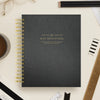 A black textured spiral-bound notebook with gold lettering reading 'DAY DESIGNER.' lies on a light beige desk surrounded by office props including pencils, a ruler, glasses, a coffee cup, and paper clips.