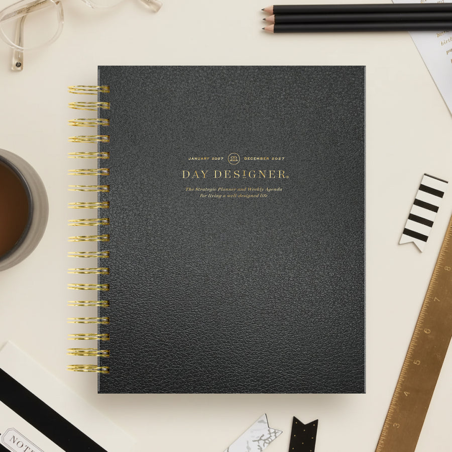 A black textured spiral-bound notebook with gold lettering reading 'DAY DESIGNER.' lies on a light beige desk surrounded by office props including pencils, a ruler, glasses, a coffee cup, and paper clips.