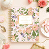A floral spiral planner titled 'DAY DESIGNER.' is arranged in a styled flat lay on a pale pink surface with roses, cookies, a teacup, and gold-toned accessories around it.