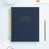 A navy blue spiral-bound notebook with gold wire binding and gold foil text reading 'DAY DESIGNER.' is shown on a light blue background, styled with a gold pen, a small card, and a decorative stem.