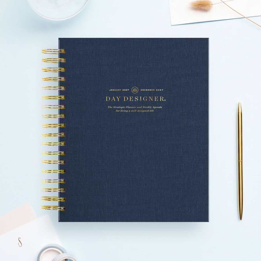 A navy blue spiral-bound notebook with gold wire binding and gold foil text reading 'DAY DESIGNER.' is shown on a light blue background, styled with a gold pen, a small card, and a decorative stem.