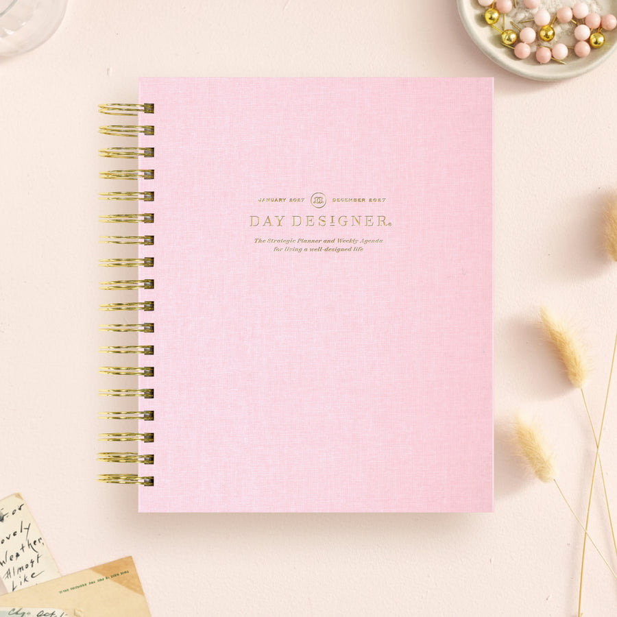 A light pink spiral-bound notebook with gold wire binding and gold foil text reading 'DAY DESIGNER.' on the cover, styled on a pale pink surface with decorative dried grass, a small dish of beads, and paper notes around it.