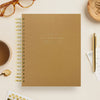 Brown notebook with gold spiral binding on a light surface with stationery items.