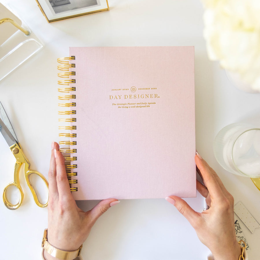 A top-down view of a person holding a light pink spiral-bound planner labeled 'DAY DESIGNER' on a white desk, surrounded by gold scissors, a framed photo, a keyboard, and a glass.