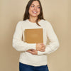 A smiling woman with long brown hair stands against a solid beige background, wearing a cream-colored ribbed knit sweater and blue jeans while holding a brown spiral-bound notebook.