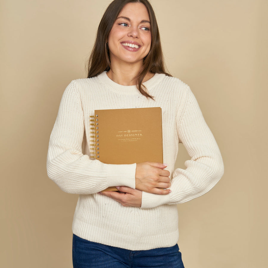 A smiling woman with long brown hair stands against a solid beige background, wearing a cream-colored ribbed knit sweater and blue jeans while holding a brown spiral-bound notebook.