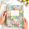 A hands-on view of a colorful spiral-bound planner or journal titled 'DAY DESIGNER.' held by two hands over a white marble surface with a cup of tea, tulips, and a gold pen nearby.