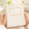 A person holds a spiral-bound notebook titled "Day Designer" with cream and gold striped cover. The scene is styled on a light tabletop with flowers, lemons, and other notebooks in the background.