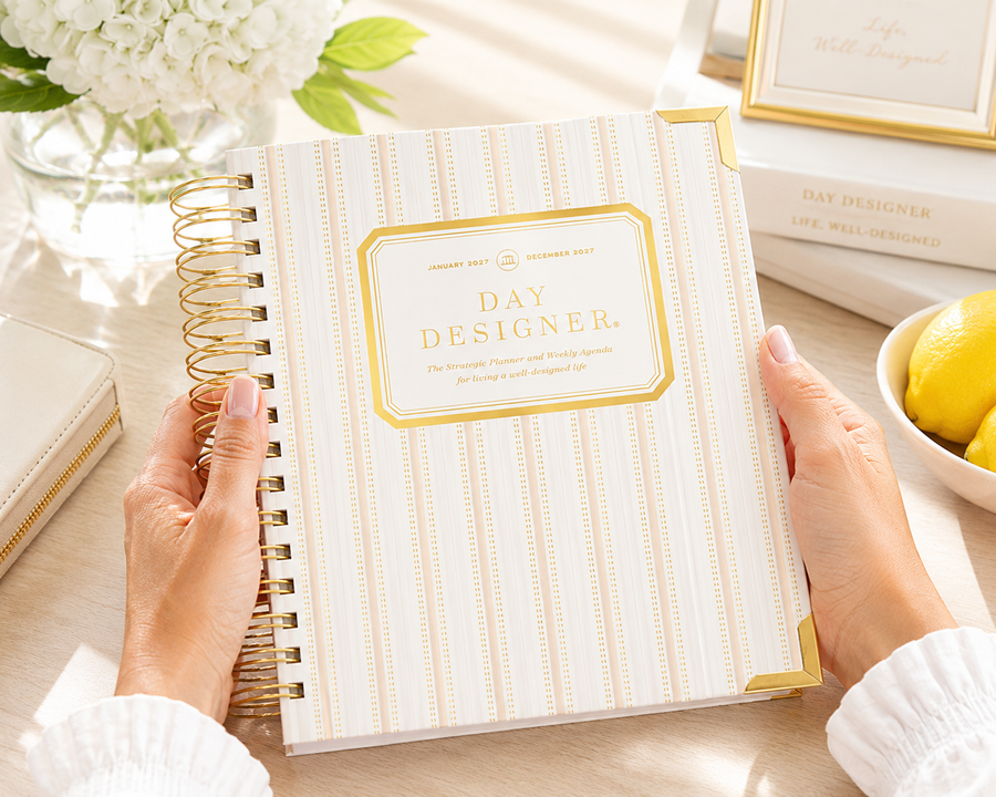 A person holds a spiral-bound notebook titled "Day Designer" with cream and gold striped cover. The scene is styled on a light tabletop with flowers, lemons, and other notebooks in the background.