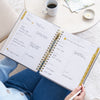 weekly open book with written on pages gold binding and tabs, hand holding white pen, blue jeans and sweater, white table and coffee cup