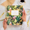 Person holding a floral notebook with a cup of coffee and decorative items on a table.