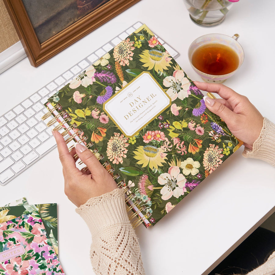 assorted flowers on deep green background, gold logo and binding, tea cup, notebooks, white keyboard and desk