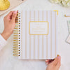 Person holding a wire-bound day designer planner with decorative cover on a marble surface with flowers and jewelry.