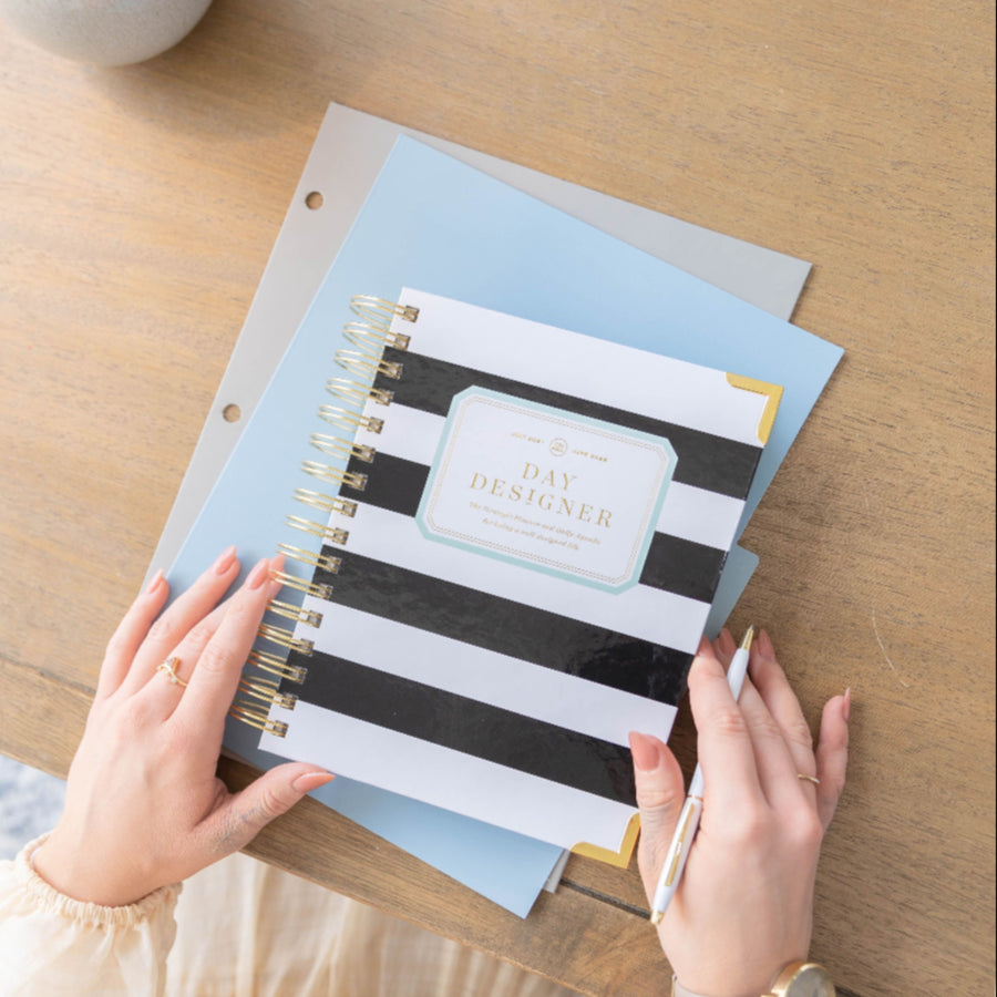 mini daily black and white stripe planner, gold day designer logo and binding, laying on light blue and clear folders, white pen, hands on wood desk