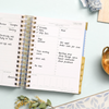 mini daily planner, open book written on pages, gold binding, tabs, and dish, dried leaves, light blue background