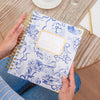 Person holding a day designer planner with repeated swan pattern on a wooden table with a cup of tea and plant in the background