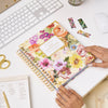 multi-colored floral planner, gold logo, binding, pen, and scissors, pink-trimmed notepad, magnetic bookmarks, white keyboard, mouse and desk