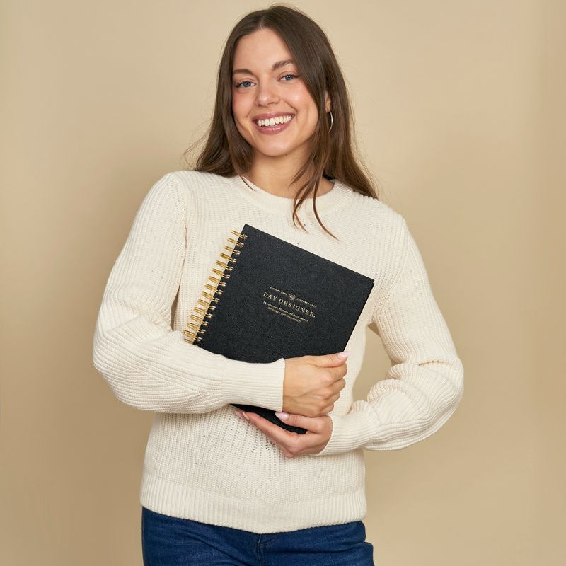 Woman holding a black planner with visible day designer branding against a beige background