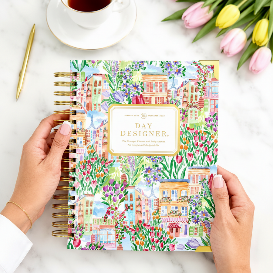 Colorful planner with floral design held by hands, next to a cup of coffee and tulips.