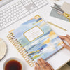 blue and gold pattern planner, gold day designer logo, binding, pen, scissors, tea cup, dish, white keyboard and desk, hands