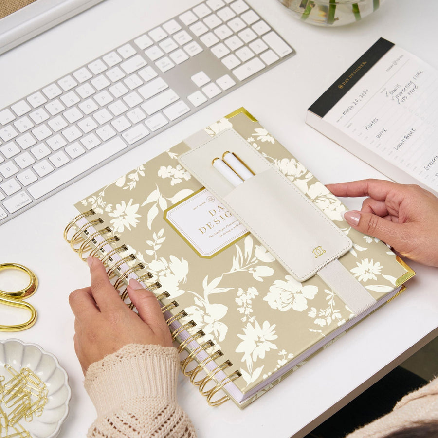 light grey planner with white flowers in silhouette pattern, elastic pen holder with two pens, black trip notepad, gold scissors and paper clips, dish, keyboard, white desk