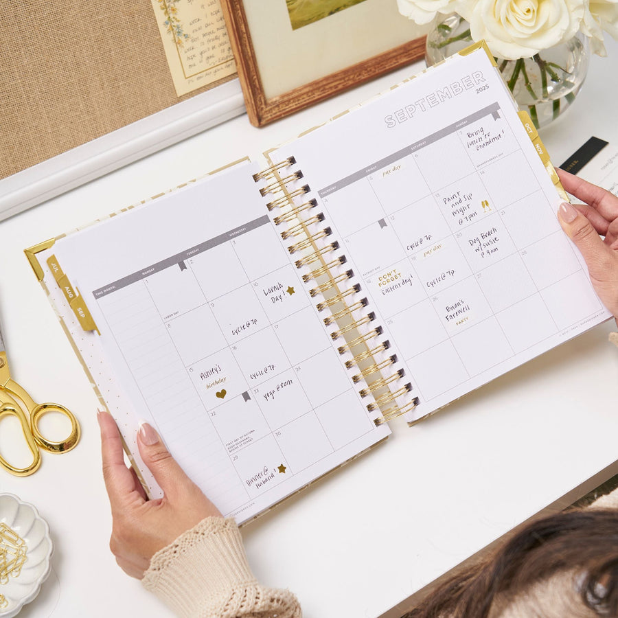 open planner, written on pages, gold scissors and paper clip, dish, notepad, flowers in vase on white desk