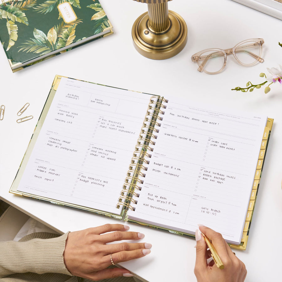 open planner book with written on pages, gold binding, tabs and paper clips, floral stem, eye glasses, brass lamp base, green and gold palm notebook on dark green background