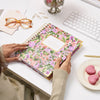 multi-colored floral planner, gold binding, pink hearts on dish, eye glasses, paper clips and pen, stationery, vase, keyboard on white desk