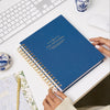 navy bookcloth planner, gold day designer logo, binding and pen, assorted notebooks, blue and white antique jars, white keyboard and desk, hands