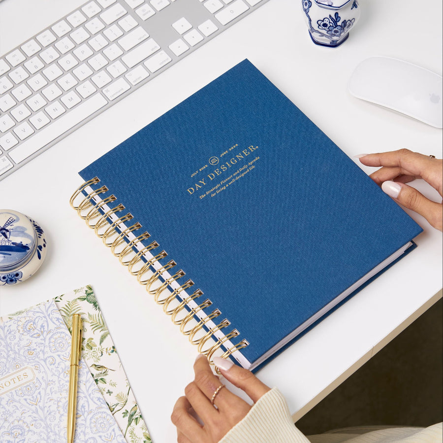 navy bookcloth planner, gold day designer logo, binding and pen, assorted notebooks, blue and white antique jars, white keyboard and desk, hands
