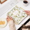 green and gold repeated pattern of birds, butterflies, fern branches, and pineapples, gold logo, binding, pen, coffee, dish with cookies, white keyboard and desk