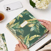 green and gold notebook on dark green background, hand, white flowers and phone, tea cup, two stacked notebooks